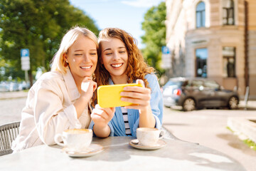 Two smiling woman are sitting in a cafe, drinking coffee, taking selfies, talking with friends. Fashion, beauty, blogging, tourism.