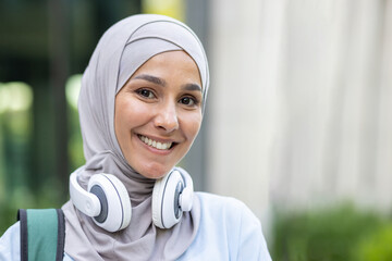 Portrait of a smiling young Muslim woman wearing a hijab and headphones around her neck, expressing...