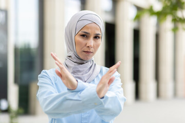 Empowered adult Muslim woman wearing a hijab crosses her arms in an 'X' gesture, signaling stop or...