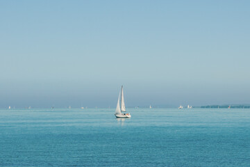 Obraz premium Small white sailboat in blue lake with many other boats far in background. Sailing yacht with one mast on a sunny day in big lake. Blue water and blue sky. Calm water without waves.