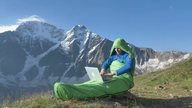 An individual, bundled in a sleeping bag, works on a laptop against the backdrop of majestic snow-capped mountains, embodying the ultimate remote work setting. A funny man defiantly works at a laptop