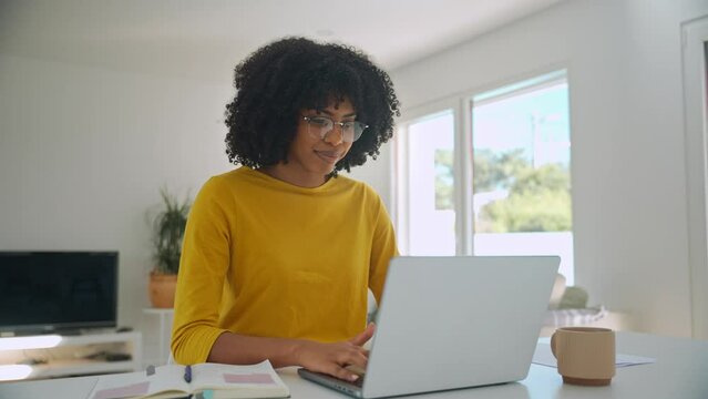 Young pretty happy smiling African American woman wearing eyeglasses using laptop computer sitting at home table hybrid working online, female student elearning, browsing web typing at pc.