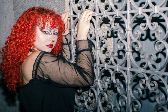Gothic curly redhead poses looking back on 
a gothic window grill