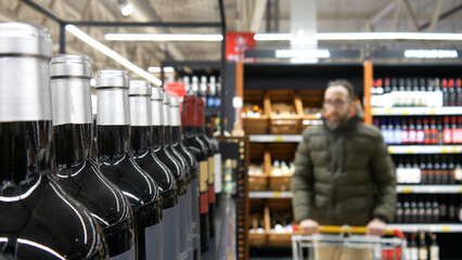 Close-up of many glass bottles of red wine in a wine shop and a man with a shopping trolley behind them