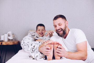 A child with bare feet is lying in bed. Dad tickles his son. Tickling the heels. Cheerful and...