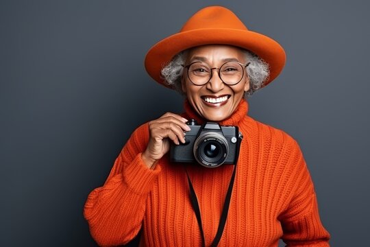 African old woman in an orange hat with brim, sweater and glasses holds camera on gray background
