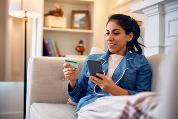 Happy Hispanic woman buying online with credit card and cell phone while relaxing at home.