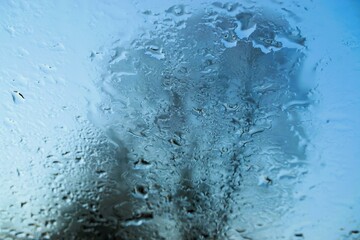 Abstraktes Motiv mit Eis- und Wassermuster auf beschlagener Glasscheibe vor hohem Baum und blauem Himmel mit Licht bei Frost, Kälte und Sonne am Morgen im Winter