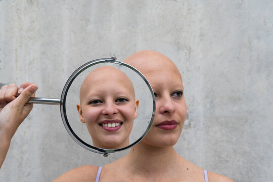 Pensive hairless woman holding mirror near face showing differen