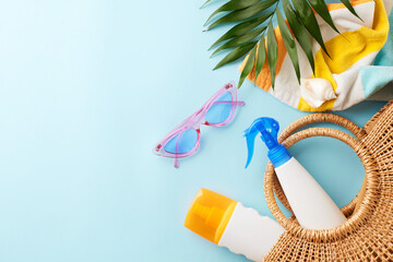 Beach day essentials: protect your skin under the sun. Top view shot of sunscreen spray, sunglasses, towel, tropical leaves, seashell, and a wicker bag on light blue background with space for messages