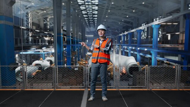 Full Body Of Asian Male Engineer With Safety Helmet Standing In Factory Manufacture of Wind Turbines. Smiling And Pointing To Side Recommends About Something While Robotic Arm Working