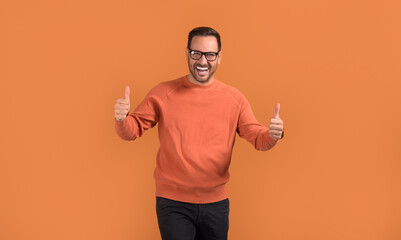 Portrait of successful young man in eyeglasses showing thumbs up signs on isolated orange background