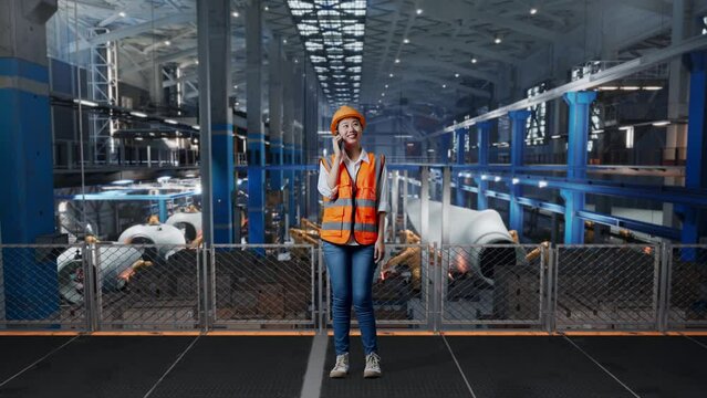 Full Body Of Asian Female Engineer With Safety Helmet Standing In Factory Manufacture Of Wind Turbines. Talking On Smartphone And Looking Around While Robotic Arm Working