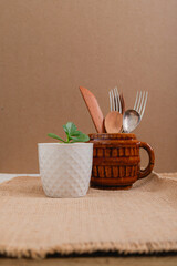 ceramic mug with kitchen cuttlery and small pot with plant on vintage tablecloth and brown wall with space for text