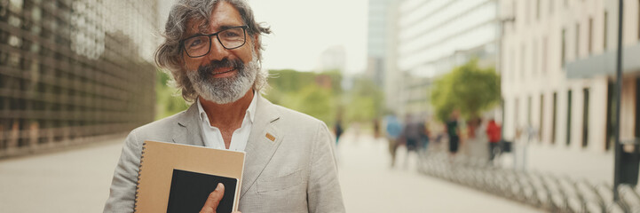 Smiling mature businessman with beard in eyeglasses wearing gray jacket looks around and walks down...