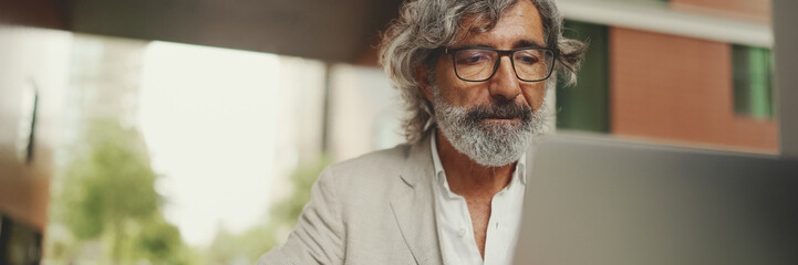 Panorama of mature businessman with beard in eyeglasses wearing gray jacket sits on cafe. Middle...
