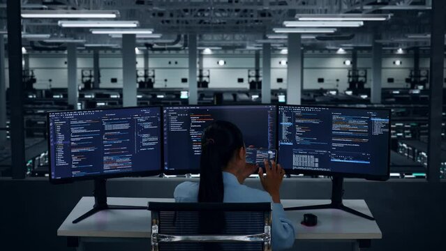 Back View Of Asian Woman Developer Working With Mobile Phone While Write Code With Multiple Computer Screens In Data Center