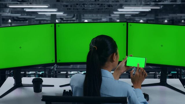 Back View Of Asian Woman Using Mobile Phone And Multiple Computer Monitor With Mock Up Green Screen In Data Center