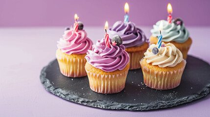 Cupcakes with colorful icing and lit candles, arranged on a slate serving plate on a purple background.