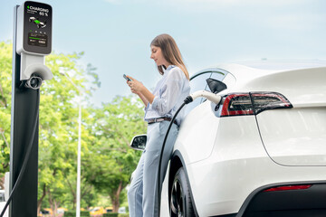 Young woman recharge EV electric vehicle battery from EV charging station and using smartphone...