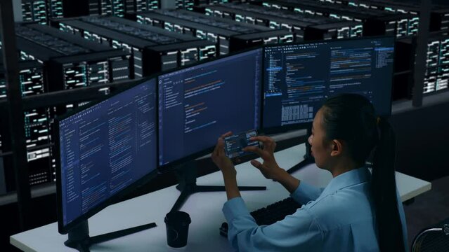 Side View Of Asian Woman Developer Working With Mobile Phone While Write Code With Multiple Computer Screens In Data Center