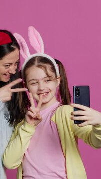 Vertical Video Playful cheery mother and girl smiling for photos on smartphone, having fun embracing each other in studio. Little child an her mom posing for pictures, fooling around. Camera A.