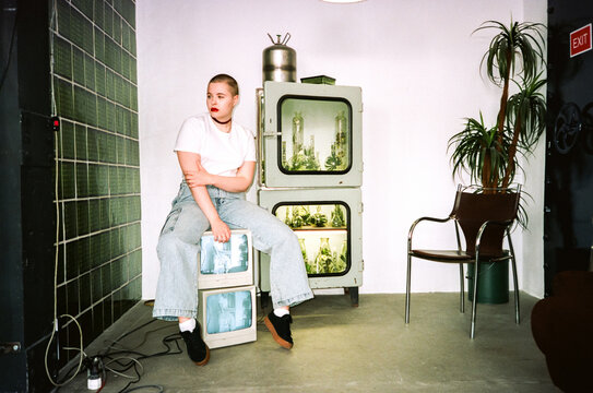 Young informal female sitting on retro TVs in creative studio