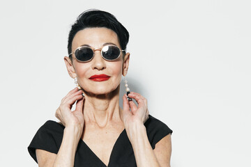 Stylish senior woman in sunglasses and earrings posing for camera against white wall background
