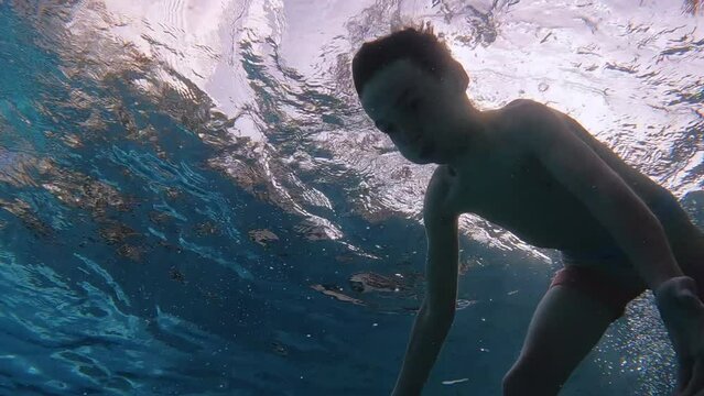 Boy takes deep breath and swims underwater in outdoor pool - from below view