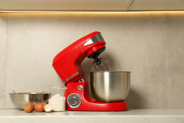 Modern red stand mixer, eggs, container with flour and bowl on white marble countertop in kitchen....
