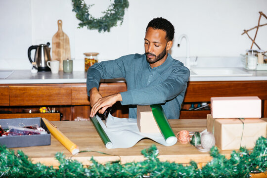 Cheerful man wrapping christmas gifts in kitchen