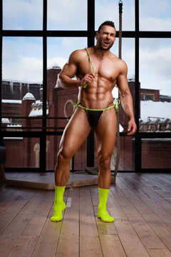 Handsome naked man in thong posing in loft studio. Male fitness model with six pack abs wearing green socks and holding green jump rope. Muscular guy in seductive underwear with sports equipment