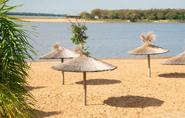 Sombrillas de paja dispersas en la playa de río al mediodía - obrazy, fototapety, plakaty