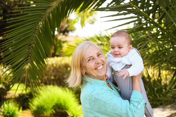 Beautiful happy smiling senior elderly woman holding on hands cute little baby boy. Grandmother and grandson having fun time together at tropical summer day in park. Multigenerational family concept