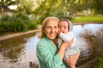 Beautiful happy smiling senior elderly woman holding on hands cute little baby boy. Grandmother and grandson having fun time together at tropical summer day in park. Multigenerational family concept