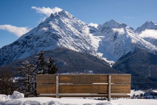 Alpine mountain panorama in winter.  The wooden backrest of a beautifully situated bench in the Engadin, Switzerland.  Snow-capped mountain peaks and white clouds against a blue sky