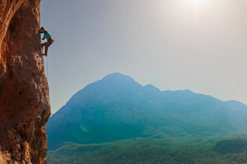 girl climber climbs a rock against a background of forest and mountains. rock climber resting on a...