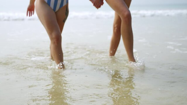 Slim feet of two young women going, making splashes by sea water. Close-up of female fitted legs walking along the sea. Two attractive girls holding hands, go at ocean water. Slow motion.