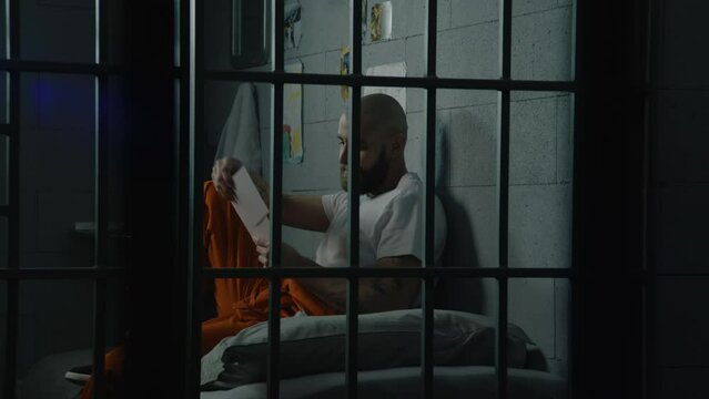 Upset male prisoner in uniform looks at pictures of family and kids sitting on bed in prison cell. Illegally convicted man serves imprisonment term in jail. Detention center or correctional facility.