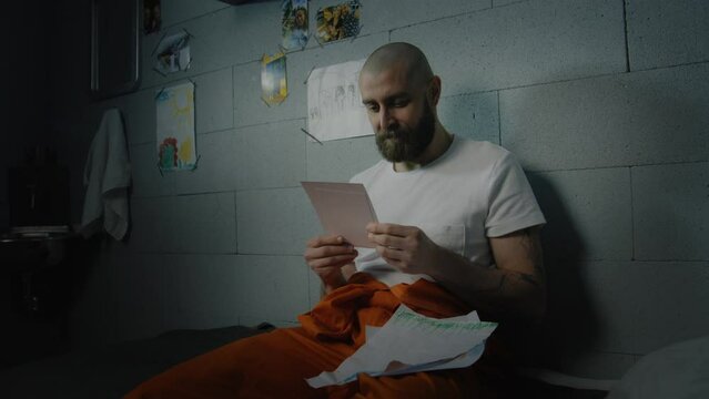 Upset male prisoner looks at family photos and kids drawings sitting on bed in prison cell. Criminal serves imprisonment term for crime in jail. Detention center or correctional facility. Portrait.
