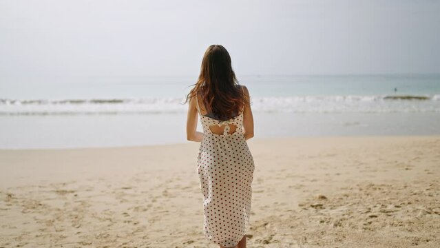 Happy young woman taking dress off and running into sea. Slim smiling brunette runs, turns in sea water waves. Fitted girl in bikini having fun, enjoying summer vacation at the ocean surf.