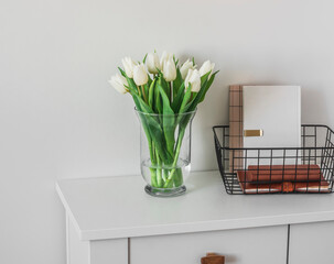 A bouquet of tulips in a glass vase, a metal basket with notebooks, books on a white wooden chest of drawers. Minimalism style