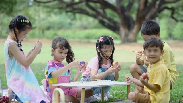 Happy group children in park, cute Asian girl and boy with mix race friend, paint egg with paintbrush together on green grass in garden. Kids celebrate Easter holiday outdoor