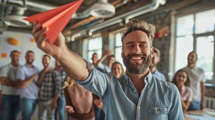 Happy male leader of company throwing red paper plane in office with group of employees smiling and talking in background. - obrazy, fototapety, plakaty