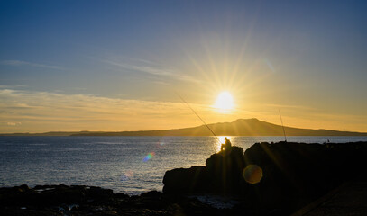 Silhouette image of a man fishing on the rocks. Sun rising over Rangitoto Island. Takapuna Beach....