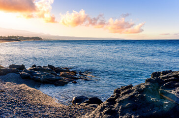 black rock sea beach during sunrise or sunset with golden sand, mild surf, blue calm water and beautiful cloudy sky with coast and mountains on background