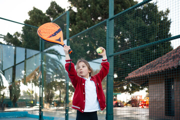 An action shot of a child reaching high to serve a padel tennis ball on an outdoor court, with palm trees in the background