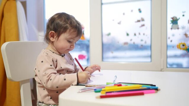 baby girl draws while sitting at a table by the window at home. happy family kid concept. baby daughter learns to draw with pencils on a sheet of paper indoors. development of fine motor skills dream