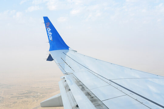 Airplane Wing Over Desert Landscape During Flight. View of a blue airplane wing with the logo Flydubai against a hazy desert backdrop from an airplane window. Dubai, UAE - August 15, 2023