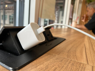 Close up the white charger connected to the pop-up electrical outlet on the wooden table in the...